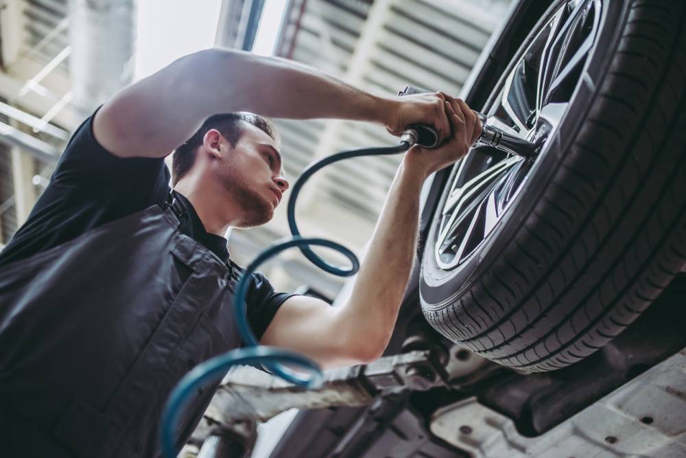 Handsome mechanic in uniform is working in auto service. Car repair and maintenance. Twisting/untwisting bolts on wheel.